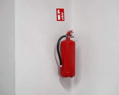 Red fire extinguisher hanged on a white wall near corner with a sticker sign